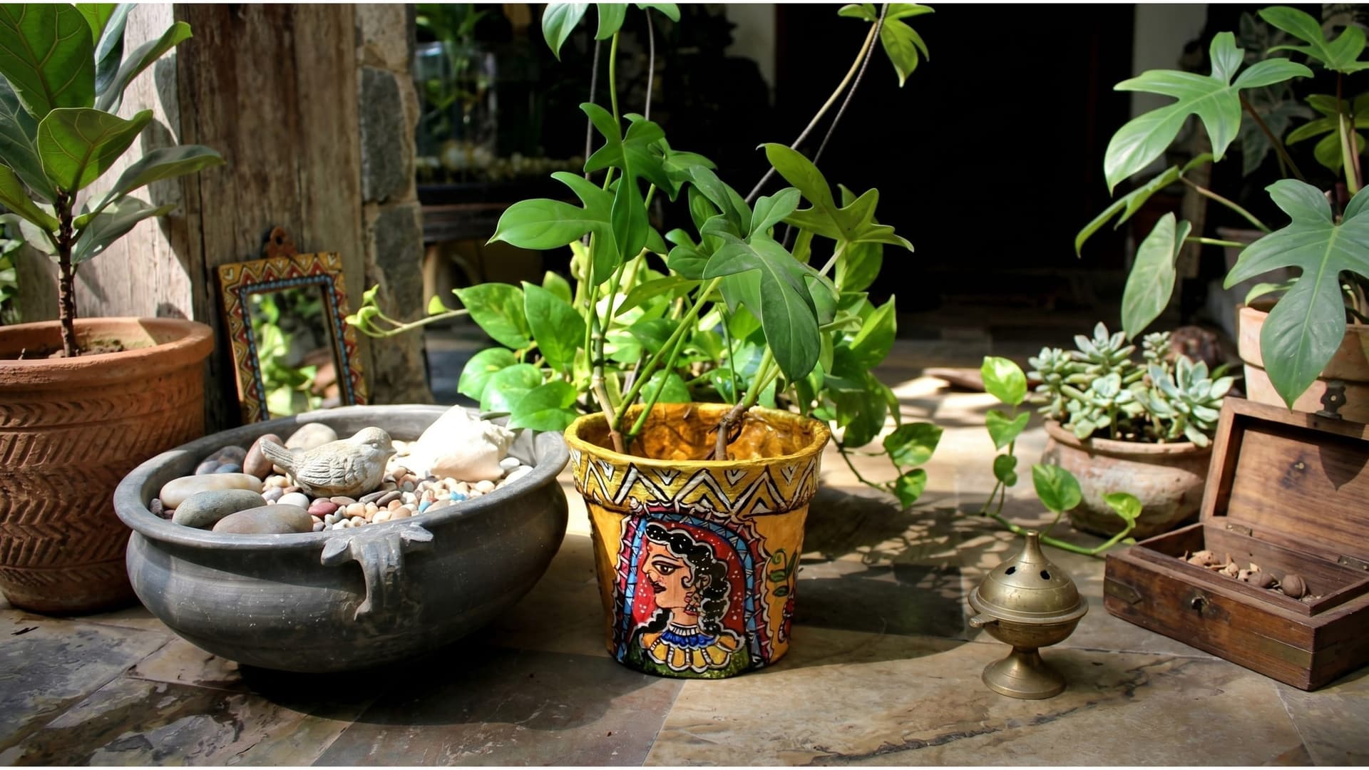 A Madhubani-painted planter in ochre, a woman's face framed by a red arch, holding a philodendron on a sunlit stone courtyard beside a stone bowl of pebbles.