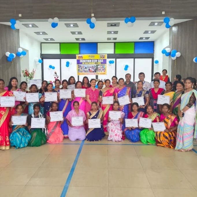 The full training group holding their certificates at the closing ceremony, beneath an exhibition-cum-sale banner.