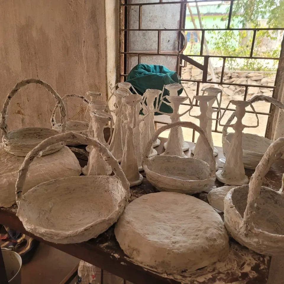 Unpainted paper-clay baskets, bowls and figurines drying on a workbench beside a barred window.