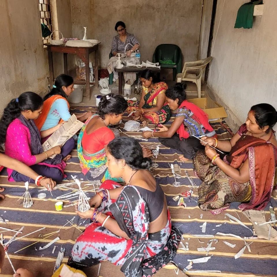 Women seated on the floor of a village room, cutting and shaping newspaper strips into paper-clay pieces.